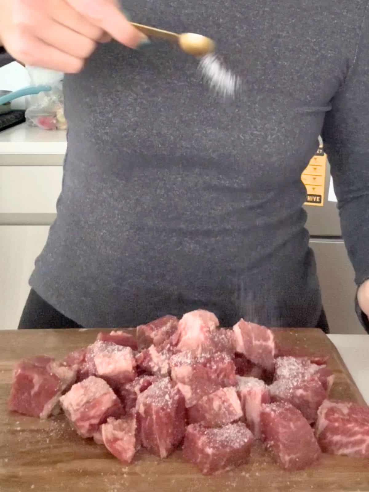 Seasoning cubed chuck beef with salt and pepper on a cutting board.