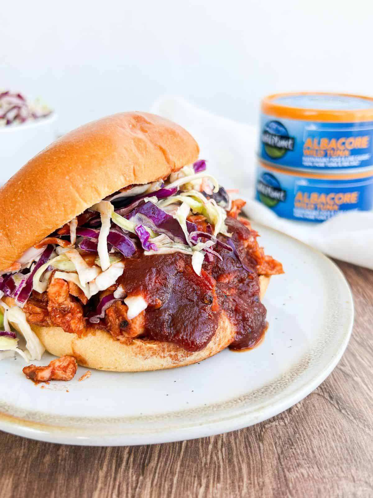 Side view of a tuna BBQ sandwich on a plate with cans of tuna in the background.
