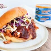 Side view of a BBQ tuna sandwich on a white plate with canned tuna in the background.