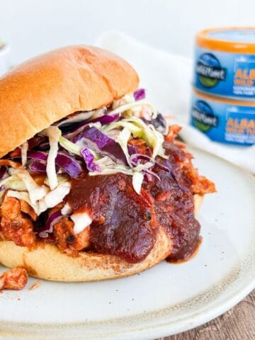 Side view of a BBQ tuna sandwich on a white plate with canned tuna in the background.