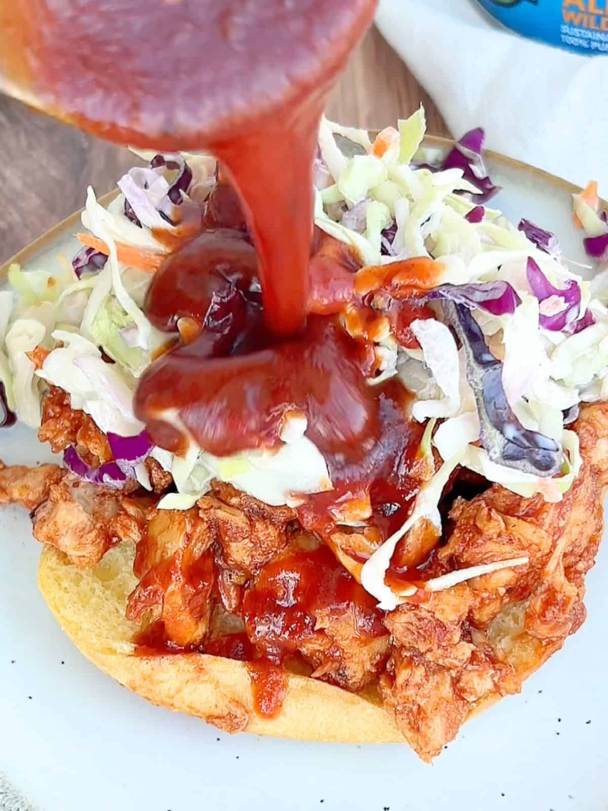 Pouring more BBQ sauce on top of the tuna and coleslaw layers on a bun.