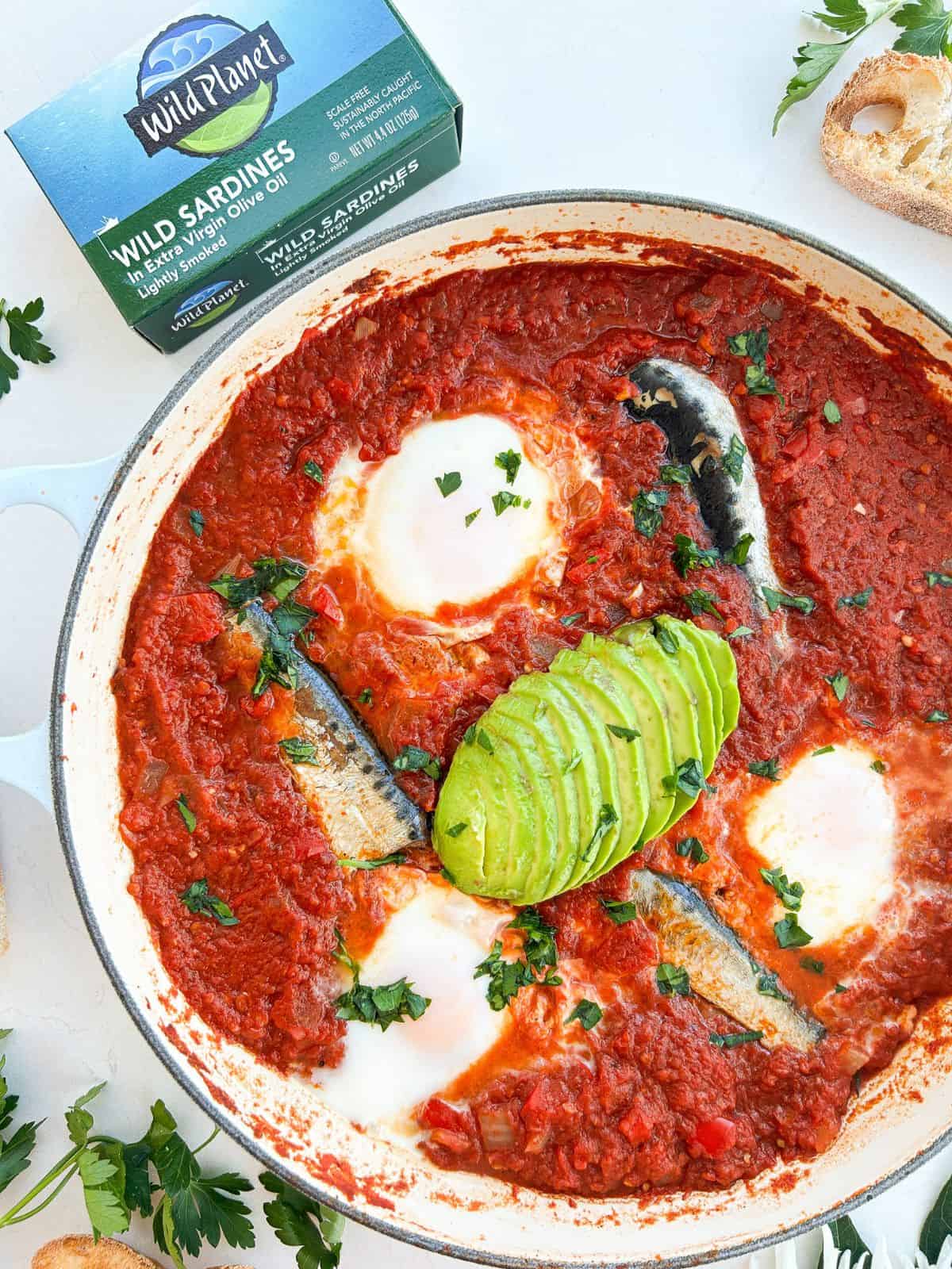 Sardine shakshuka in a skillet garnished with avocado and parsley.