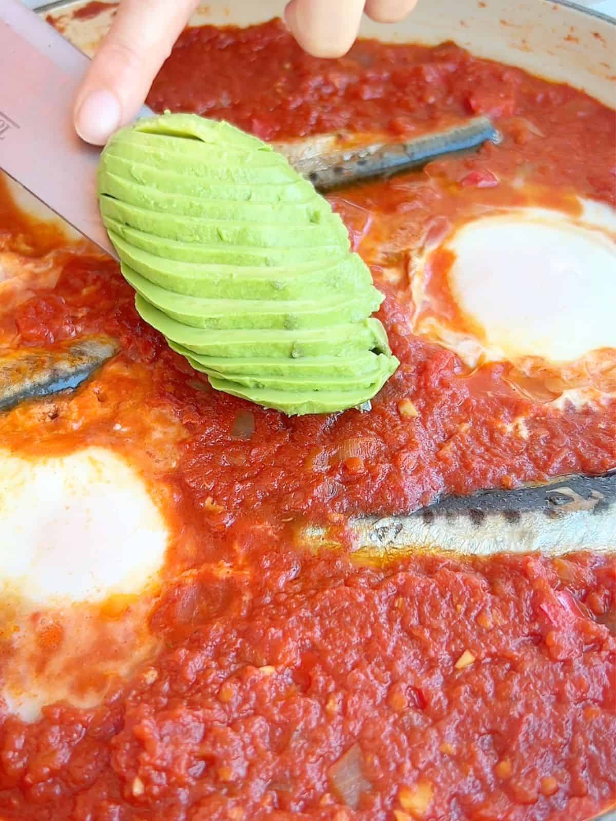 Placing a sliced avocado on the finished sardine shakshuka.