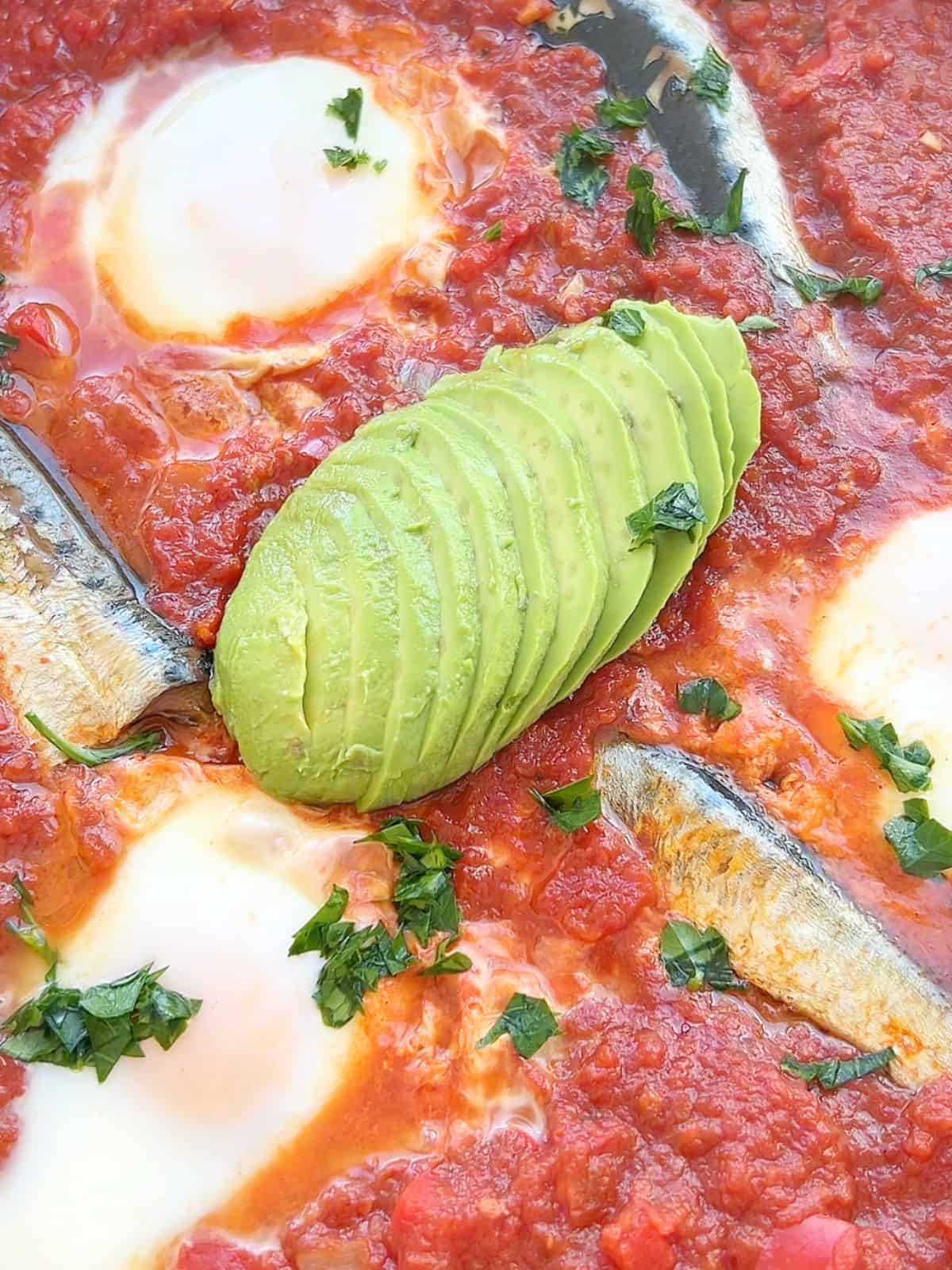 Close up of sardine shakshuka topped with avocado and parsley.