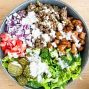 Overhead shot of the finished burger bowl with lettuce, pickles, onions, ground beef, tomatoes, potatoes and drizzled with a burger sauce.