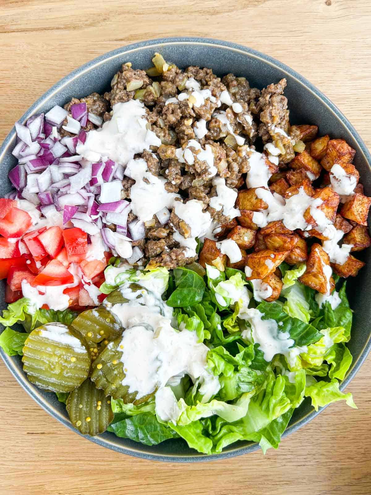 Closer shot of the finished burger bowl with lettuce, pickles, onions, ground beef, tomatoes, potatoes and drizzled with a burger sauce.