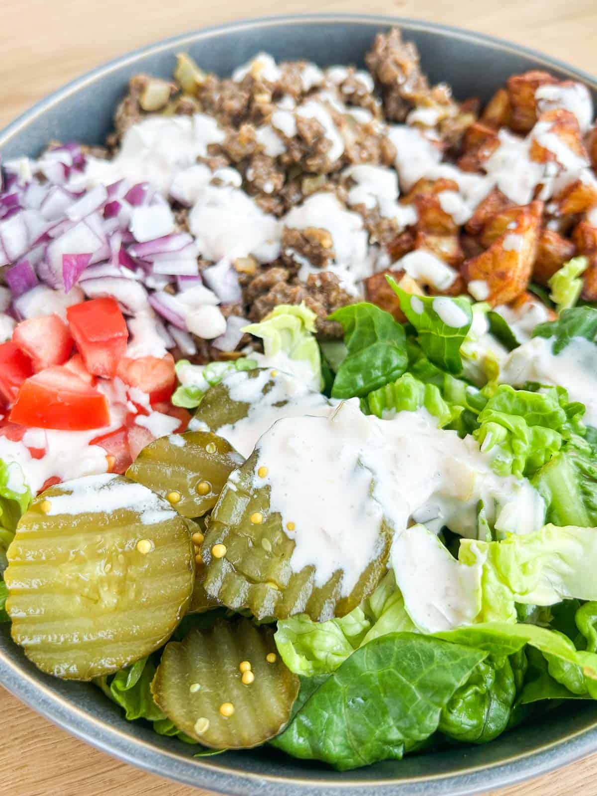 Side shot of the finished burger bowl with lettuce, pickles, onions, ground beef, tomatoes, potatoes and drizzled with a burger sauce.