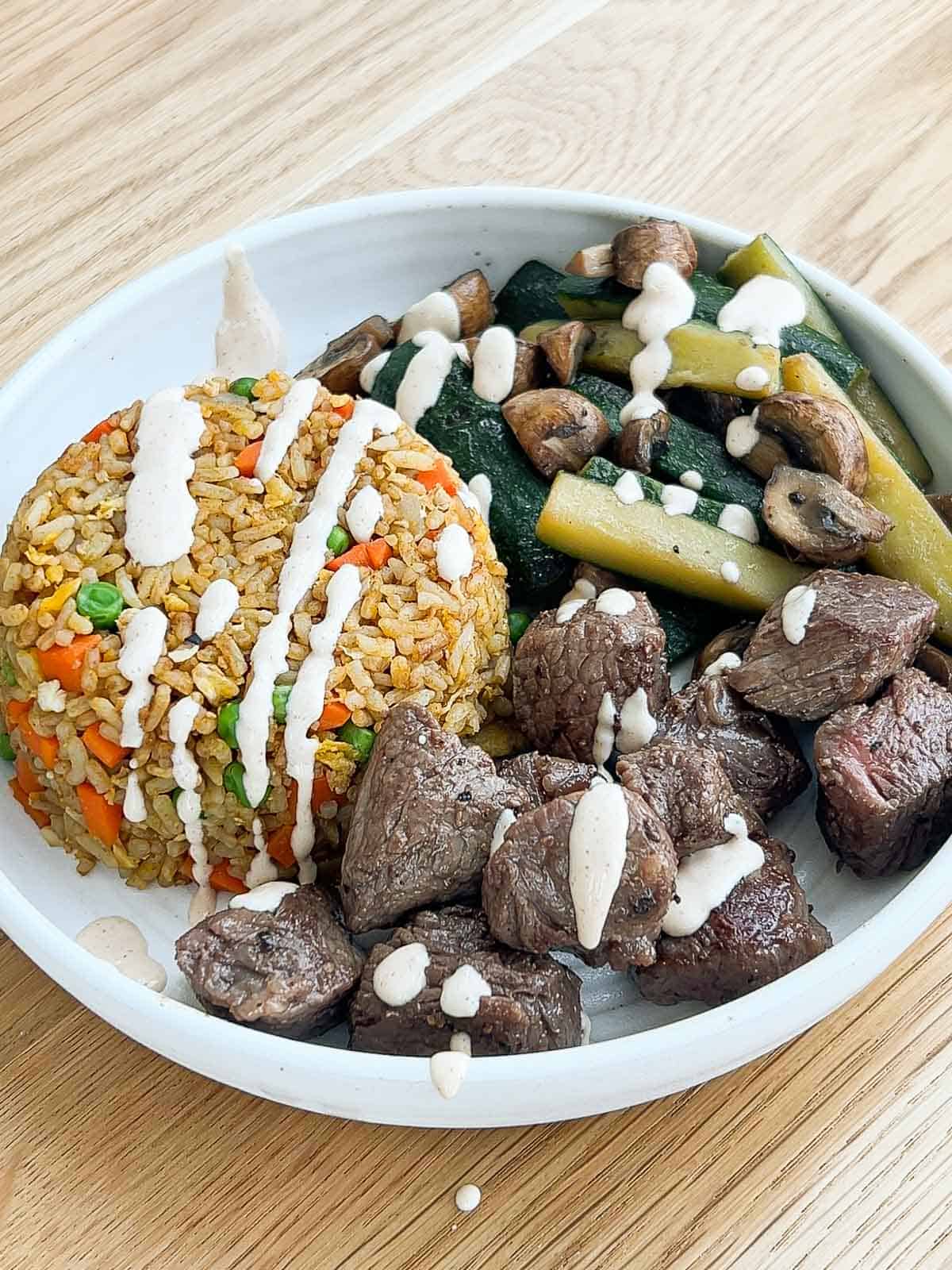 Steak, veggies, fried rice in a bowl plate drizzled with yum yum sauce.