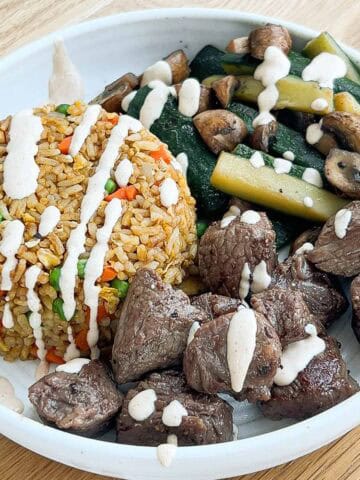 Close up shot of steak, veggies, fried rice in a bowl plate drizzled with yum yum sauce.