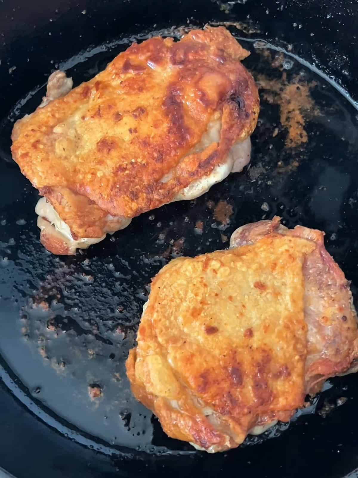 Two chicken thighs with crispy skin cooking in a cast iron pan.