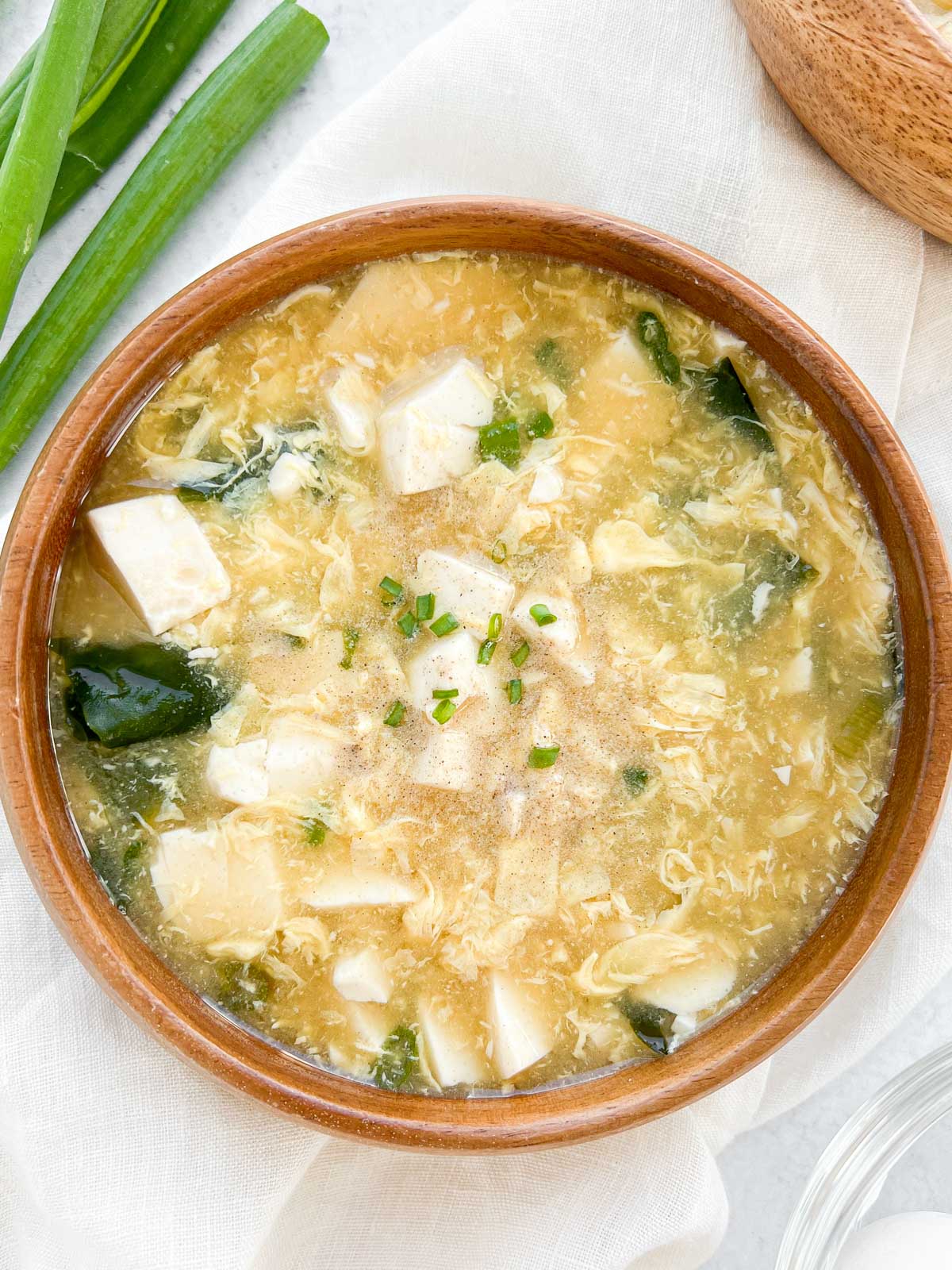 Creamy egg drop soup with tofu, green onions, and seaweed in a rustic wooden bowl. Perfect comfort food for cold days.