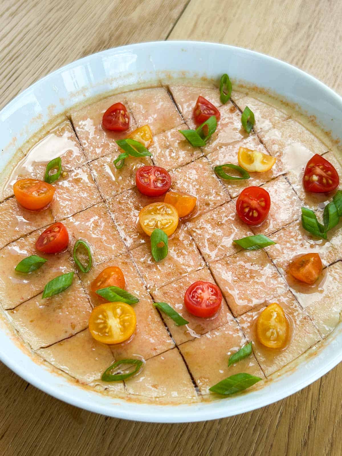 Zoomed out overhead shot of steamed egg cut into cubes and topped with scallions, cut baby tomatoes, soy sauce and sesame oil.