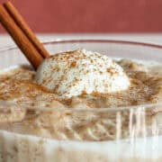Close up shot of rice pudding in a glass with a dollop of whipped cream and a cinnamon stick, sprinkled with ground cinnamon.