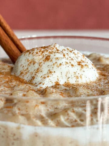 Close up shot of rice pudding in a glass with a dollop of whipped cream and a cinnamon stick, sprinkled with ground cinnamon.