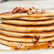 Side shot of stack of pancakes with pecan pieces on top and maple syrup dripping down the side.