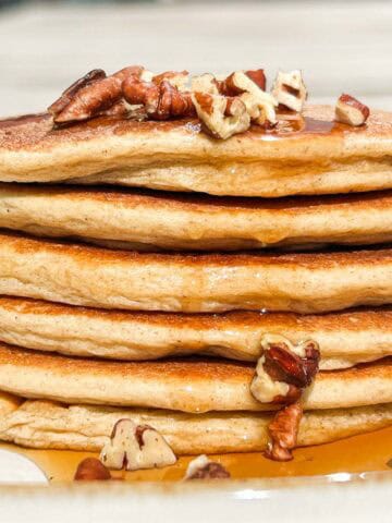 Side shot of stack of pancakes with pecan pieces on top and maple syrup dripping down the side.