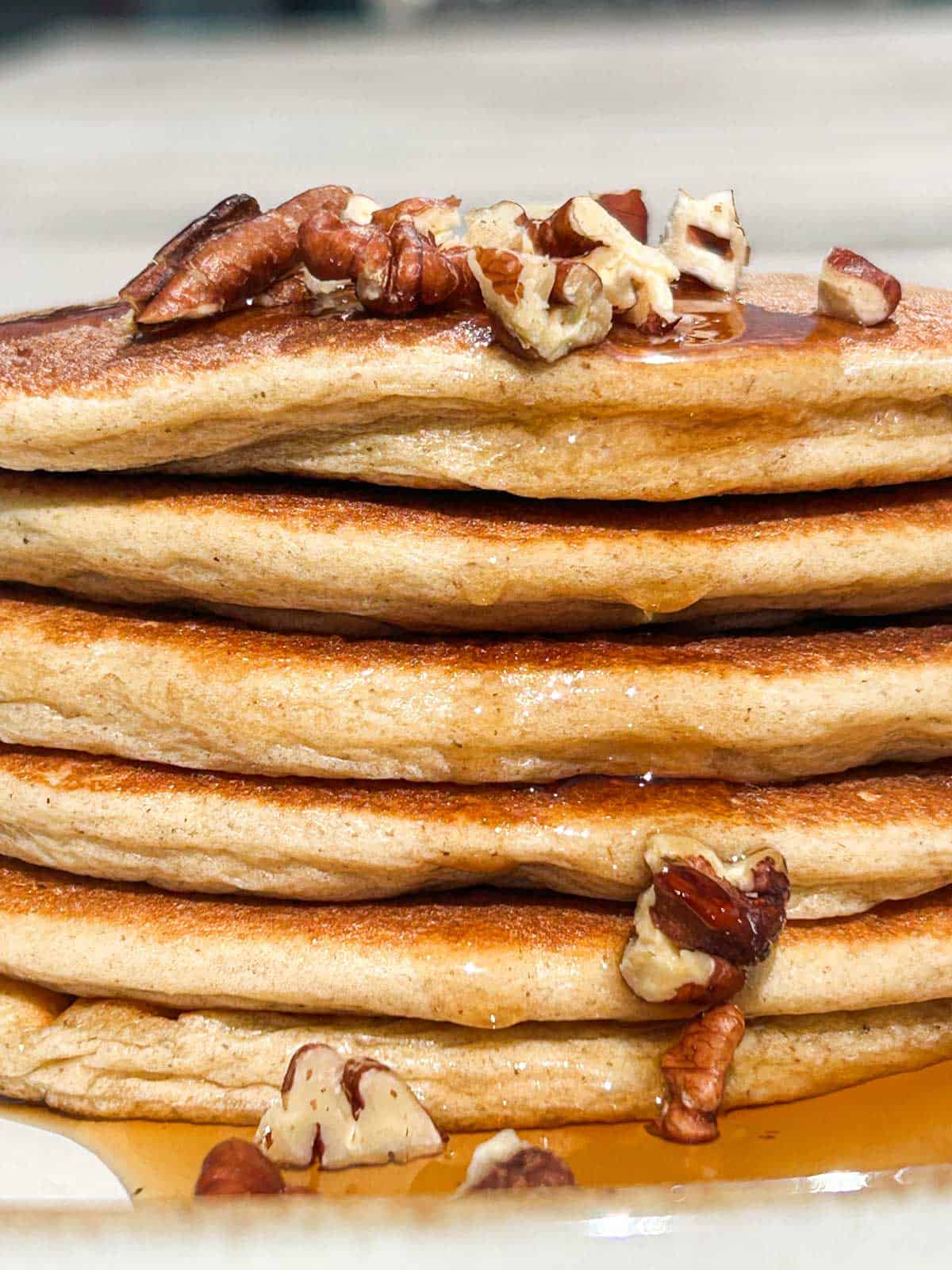 Close up shot of stack of pancakes with pecan pieces on top and maple syrup dripping down the side but longer shot.