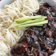 Close up of jjajangmyeon noodles and sauce in a bowl, separated, with thinly sliced cucumbers on top.