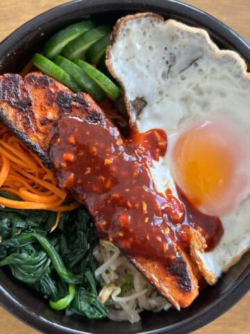 Overhead shot of salmon with gochujang sauce, spinach, carrots, soybean sprouts, cucumber and a fried egg in a 1x1 format.