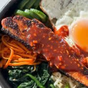 Close up shot of salmon with gochujang sauce, spinach, carrots, soybean sprouts, cucumber and a fried egg in a 1x1 format.