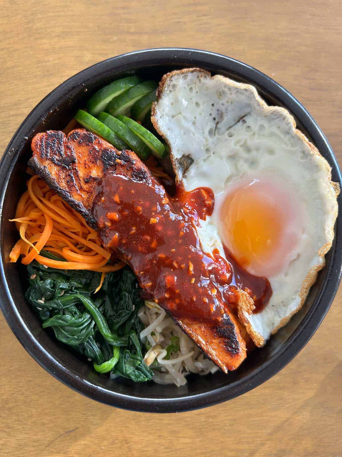 Overview shot of bibimbap bowl with carrots, soybean sprouts, fried egg, salmon, spinach and cucumber.