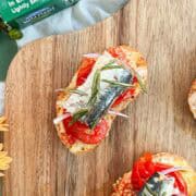 Sardines on roasted tomatoes on baguette slices on a board.