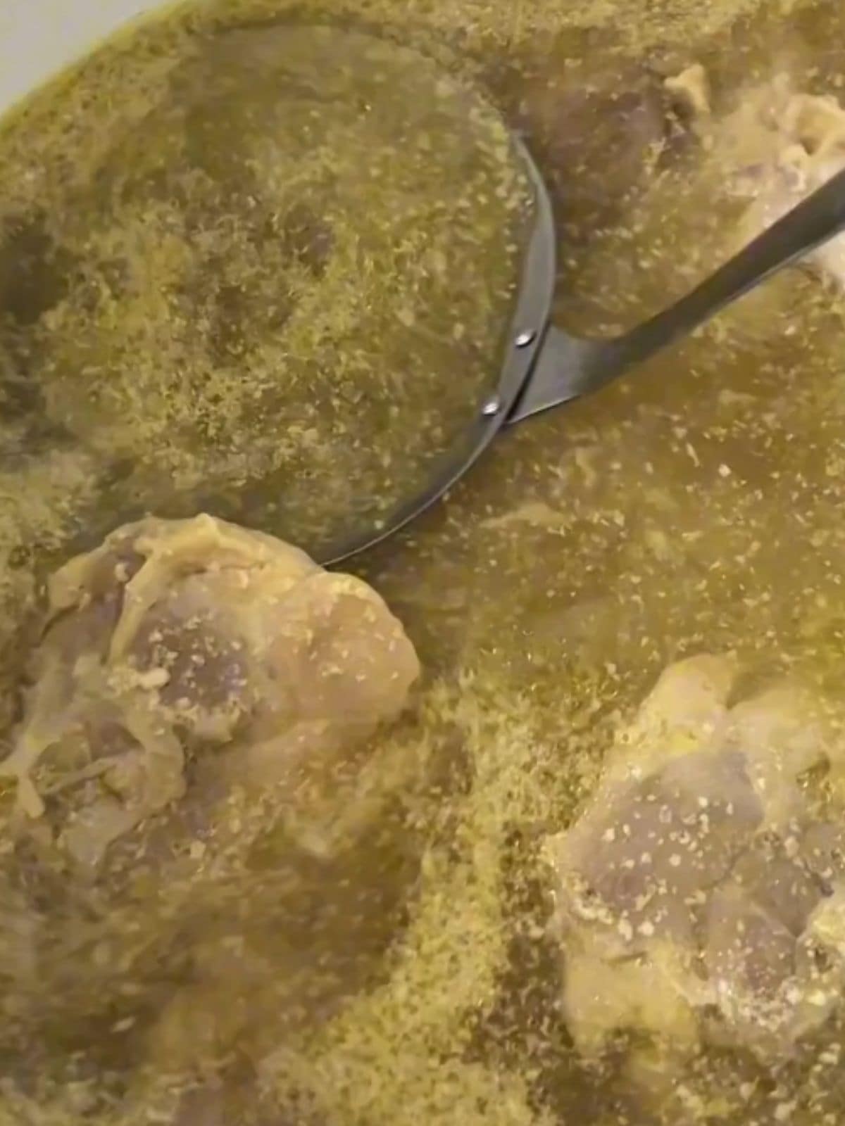 Close-up of chicken thighs simmering in broth as a ladle skims off impurities, with onion and ginger removed.