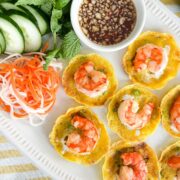 Crispy bánh khọt topped with shrimp, served with fresh herbs, cucumber, pickled vegetables, and a side of fish sauce for dipping.