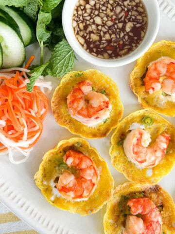 Crispy bánh khọt topped with shrimp, served with fresh herbs, cucumber, pickled vegetables, and a side of fish sauce for dipping.
