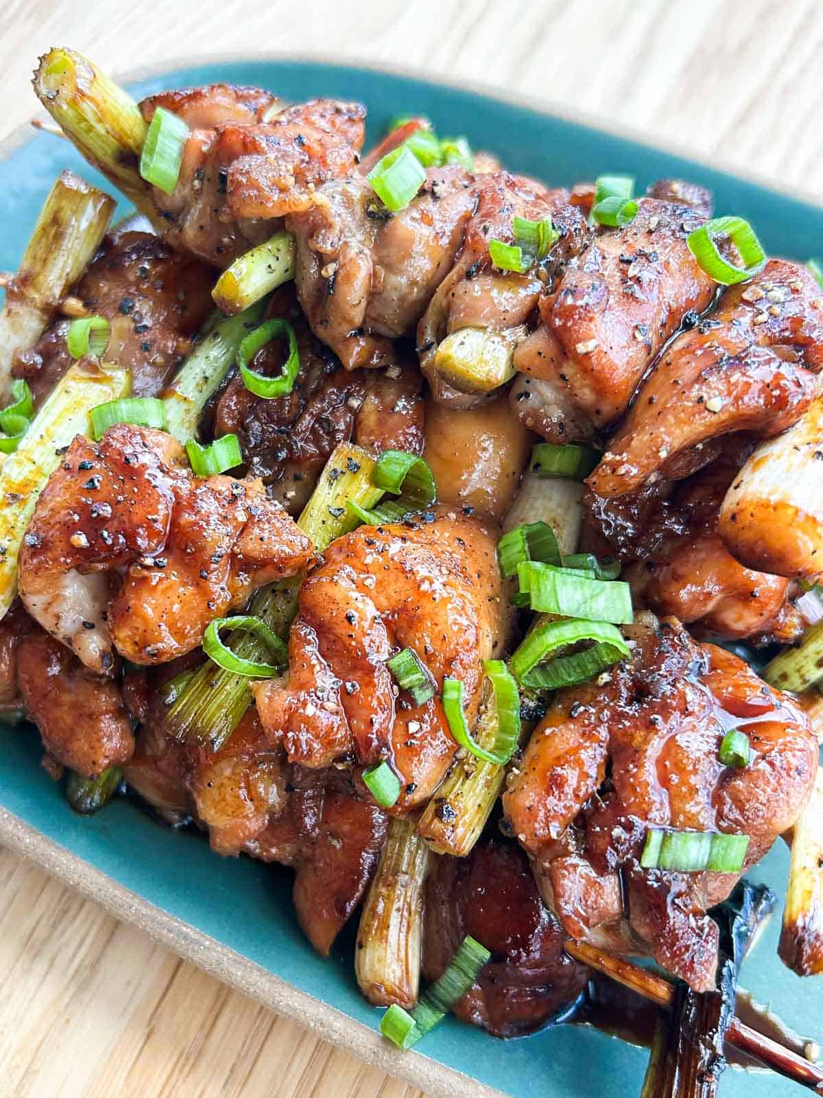 Close-up of glazed Vietnamese chicken yakitori skewers piled on a teal plate, garnished with fresh scallions and cracked black pepper.
