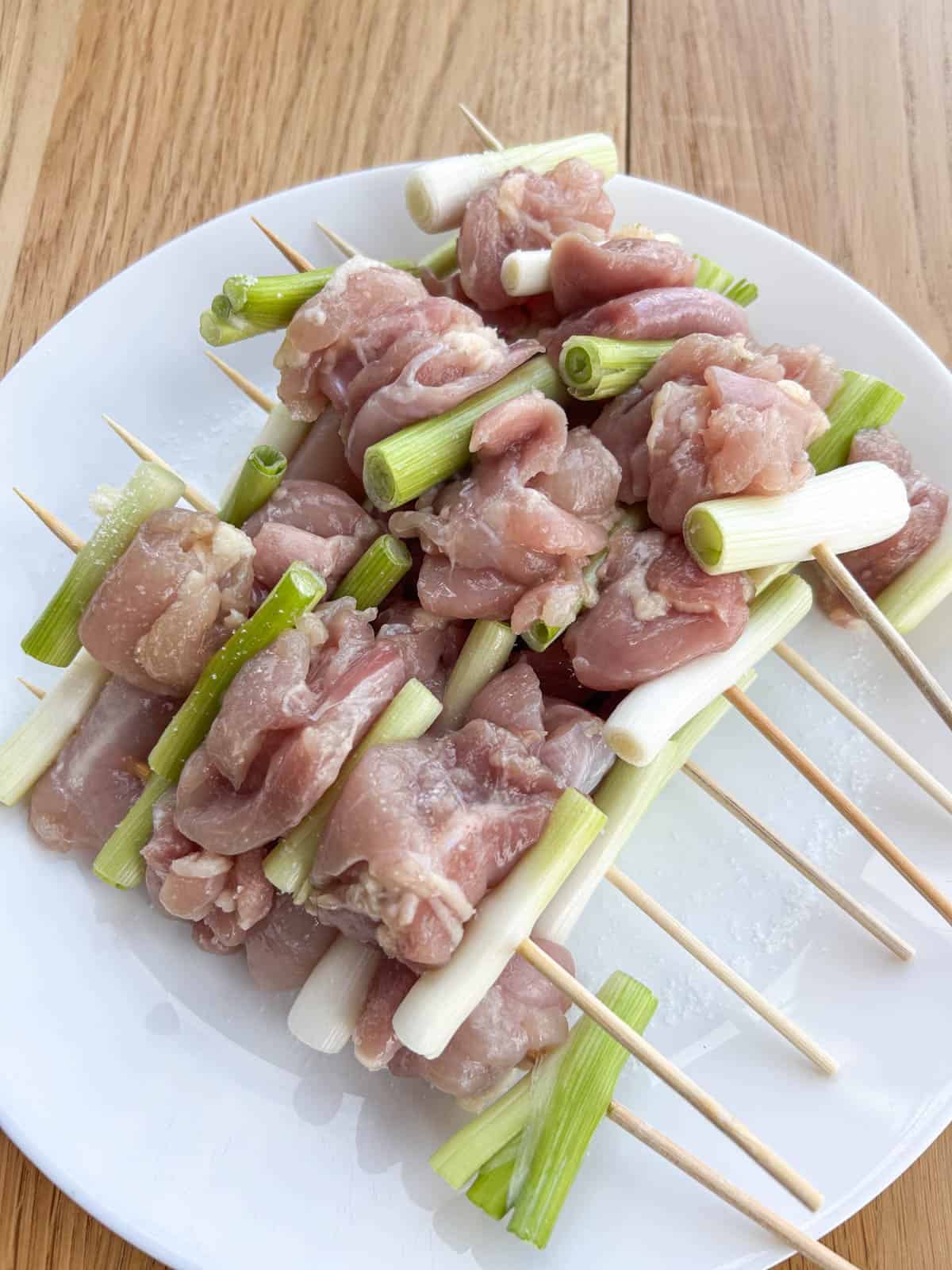 Raw chicken thigh pieces and scallions threaded onto bamboo skewers, lightly sprinkled with salt on a white plate.