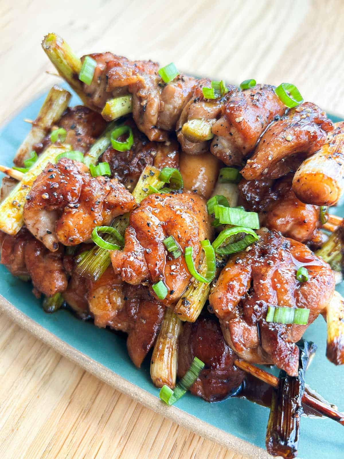 Sticky, charred Vietnamese yakitori skewers stacked on a teal plate with sliced green onions on top.