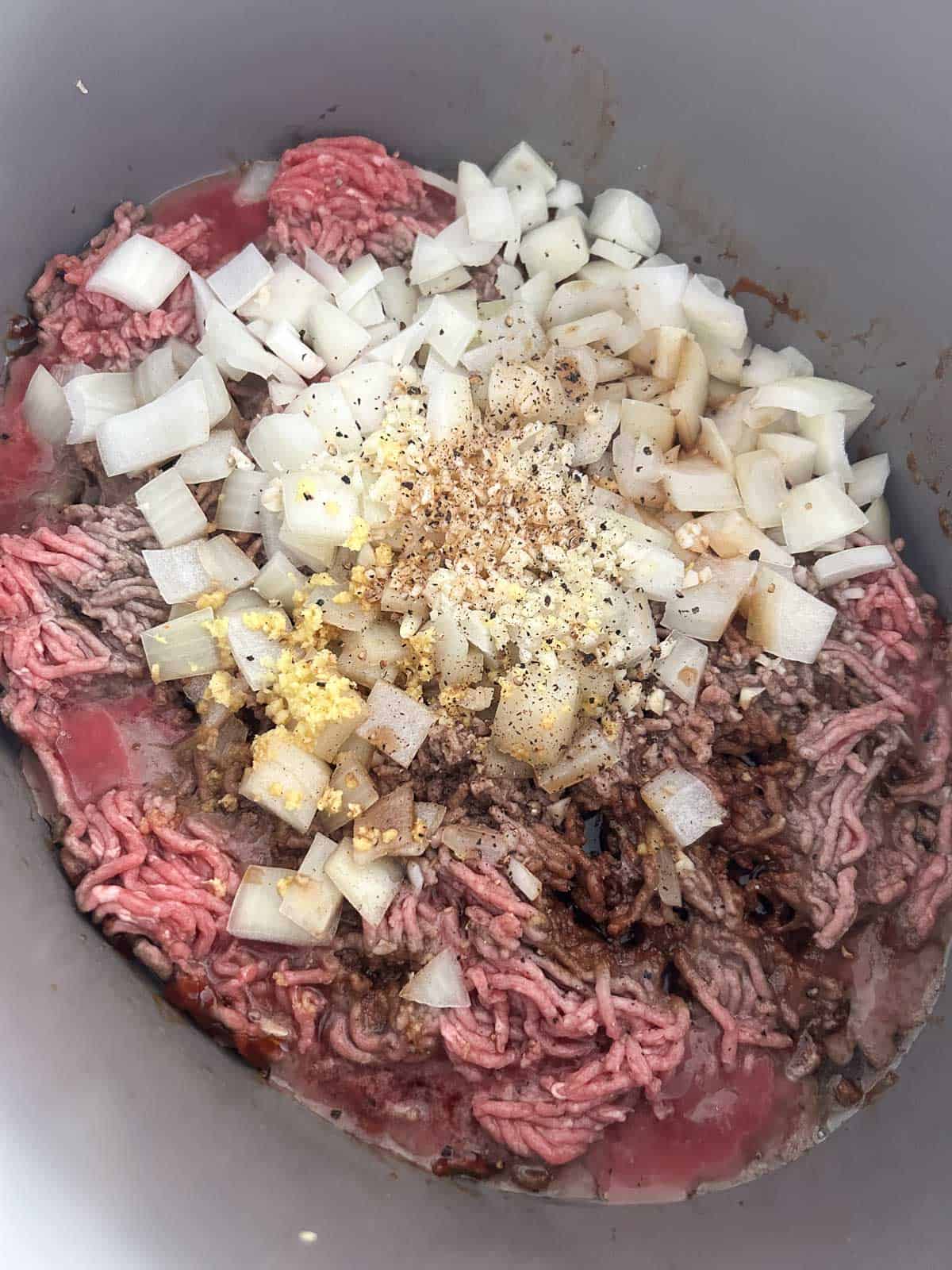 Raw ground beef topped with diced onion, minced garlic, grated ginger, and black pepper over caramel in a pot.