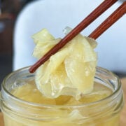 Wooden chopsticks lifting a bundle of pale yellow pickled ginger slices out of a glass jar.