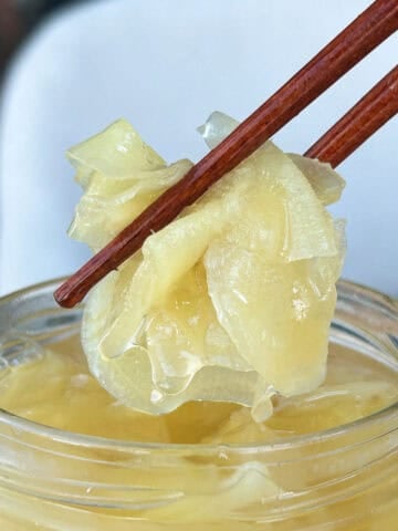 Wooden chopsticks lifting a bundle of pale yellow pickled ginger slices out of a glass jar.