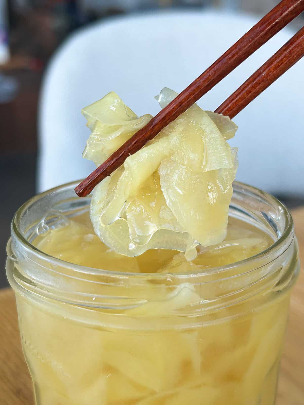 Chopsticks holding up homemade pickled ginger slices above a jar of ginger in sweet-tangy brine.